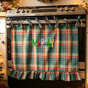 May include: A green, red, and yellow tartan oven handle towel featuring a ruffled bottom edge and fabric ties at the top. The centre of the towel displays the text Merry Christmas in colourful lettering with a small green tree graphic.