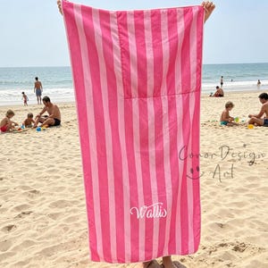 May include: A rectangular beach towel featuring vertical stripes in alternating shades of light pink and dark pink. The towel displays the white cursive text Wallis near the bottom center and includes a watermark reading Conor Design Art.