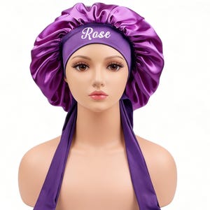 May include: A purple satin bonnet with the name "Rose" embroidered in white. The bonnet has a wide band and long ties. The bonnet is displayed on a mannequin head against a white background.