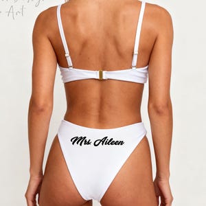 Könnte beinhalten: Weißer zweiteiliger Badeanzug mit hoch tailliertem Höschen und Oberteil mit verstellbaren Trägern. Auf der Unterseite des Badeanzugs steht in schwarzer Schrift "Mrs. Aileen". Das Oberteil hat einen goldenen Verschluss am Rücken.