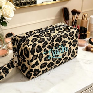 May include: A rectangular makeup bag with a black zipper and a wrist strap. The bag features a tan background with black leopard spots and the name "Lillian" embroidered in light blue. Makeup and brushes are visible on the table.
