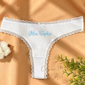 May include: White ribbed thong underwear with a delicate lace trim and a small bow. The text "Mrs. Sophie" is embroidered in light blue. A sprig of greenery and a cotton ball are in the background.