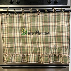 May include: A green, cream and red plaid fabric curtain with a ruffled bottom edge, hanging from a black rod. The curtain features an embroidered yellow floral sprig and the black text "The Memaw" centred on the front surface.