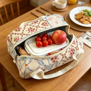Pode incluir: Uma lancheira com padrão floral com uma alça branca, aberta para revelar um recipiente branco, uma maçã vermelha e tomates cereja. A bolsa tem um design acolchoado com flores rosa e detalhes azuis. A bolsa está em uma mesa de madeira.