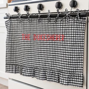 May include: A black and white gingham oven handle towel featuring a ruffled bottom edge and fabric ties. The towel is embroidered with the red text THE DUKESHERER and is draped over the handle of a white kitchen stove with five black knobs.
