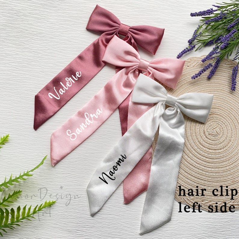 May include: Four satin hair clips in shades of pink and white, each with a bow and long streamer. The streamers are printed with names in white script: Valerie, Sandra, and Naomi. The text "hair clip left side" is also visible.