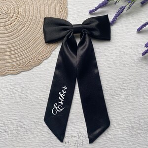 May include: A black satin bow with long, flowing tails. The name "Esther" is written in white script on one of the tails. The bow is likely for hair or as an accessory.
