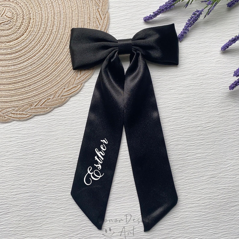 May include: A black satin bow with long, flowing tails. The name "Esther" is written in white script on one of the tails. The bow is likely for hair or as an accessory.