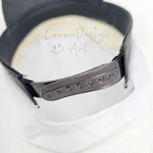 May include: A white baseball cap with a black brim and a black plastic snapback closure at the rear. The interior of the cap features the text Conor Design Art with a small smiley face icon printed in a light grey color on the white fabric.