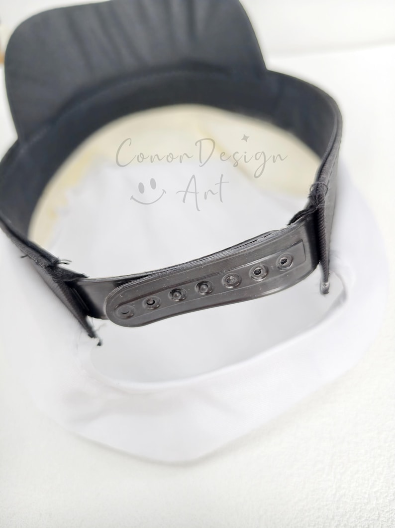 May include: A white baseball cap with a black brim and a black plastic snapback closure at the rear. The interior of the cap features the text Conor Design Art with a small smiley face icon printed in a light grey color on the white fabric.