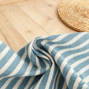 May include: Close-up of a blue and white striped fabric with the text "Conor Design Art". The fabric is folded, showing the texture and pattern. A woven circular placemat is in the background.
