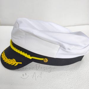 May include: A white captain's hat featuring a black brim with a yellow embroidered laurel leaf design. A yellow braided cord is attached across the front with a small gold-toned button on each side. The hat has a structured white crown.