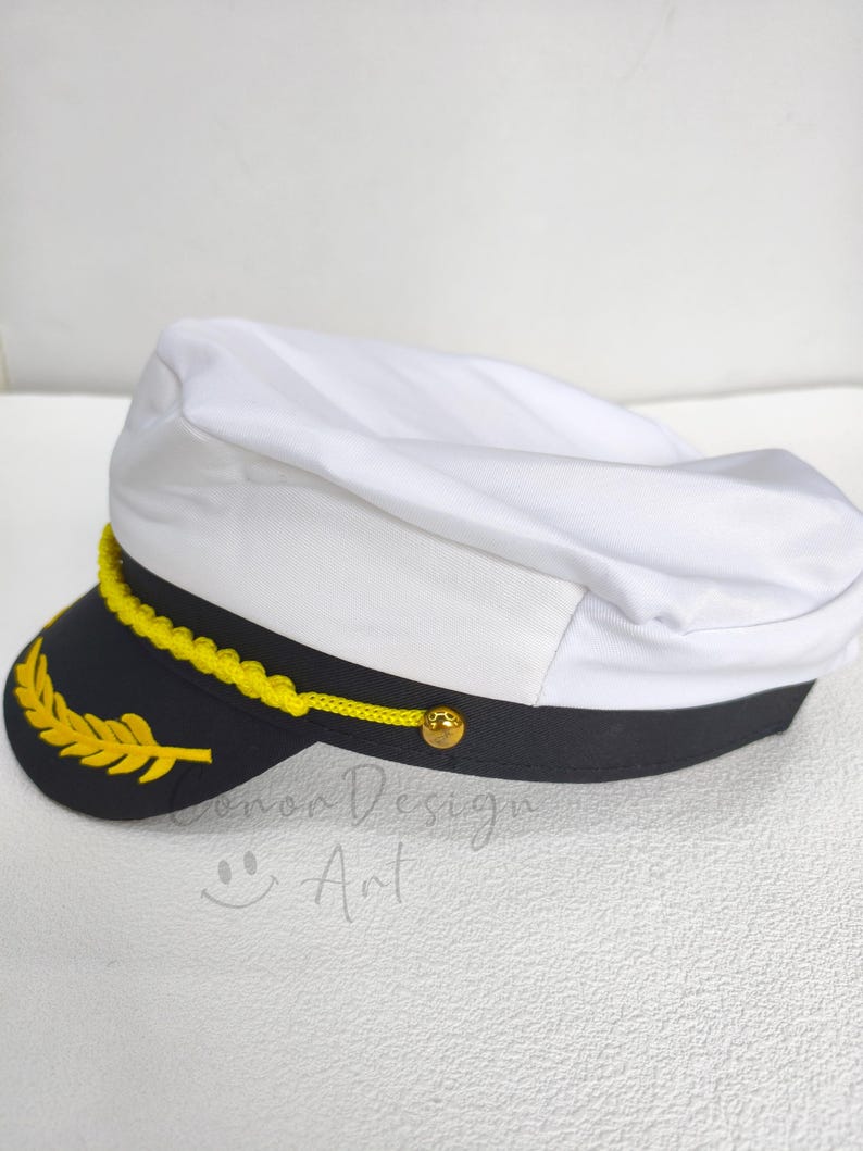 May include: A white captain's hat featuring a black brim with a yellow embroidered laurel leaf design. A yellow braided cord is attached across the front with a small gold-toned button on each side. The hat has a structured white crown.