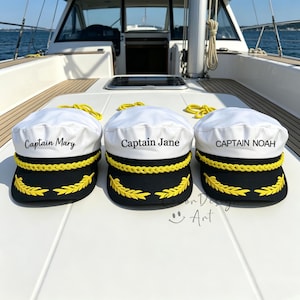 May include: Three white and black captain hats with yellow braided trim and yellow leaf embroidery on the brim. Each hat features custom embroidered text: Captain Mary, Captain Jane, and CAPTAIN NOAH, displayed on the white crown of the caps.