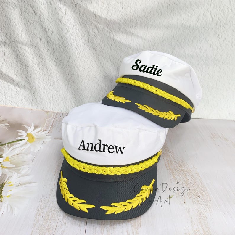 May include: Two white captain hats with dark gray brims, each featuring a yellow braided cord and yellow embroidered laurel leaf accents. The top hat is personalized with the name Sadie, and the bottom hat is personalized with the name Andrew.