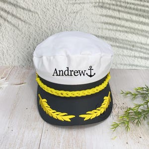 May include: A white and black captain's hat featuring the embroidered name Andrew in black lettering next to a black anchor icon. The hat includes a yellow braided cord across the front and yellow embroidered leaf detailing on the black brim.
