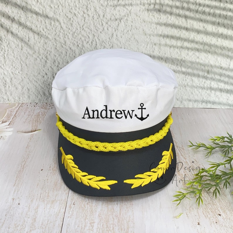 May include: A white and black captain's hat featuring the embroidered name Andrew in black lettering next to a black anchor icon. The hat includes a yellow braided cord across the front and yellow embroidered leaf detailing on the black brim.