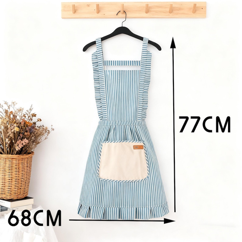 May include: A blue and white striped apron with a large cream-colored pocket. The apron is 26.77 inches wide and 30.31 inches long. It has ruffled edges and is hanging on a black hanger.