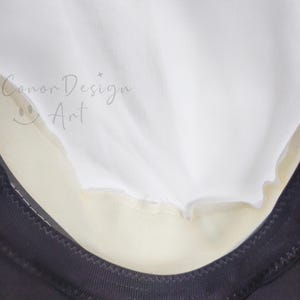 May include: A white fabric layer overlapping a cream-colored section, bordered by a black trim with visible stitching. The text 'Conor Design Art' with a small smiley face icon is printed in a light gray script on the white fabric surface.