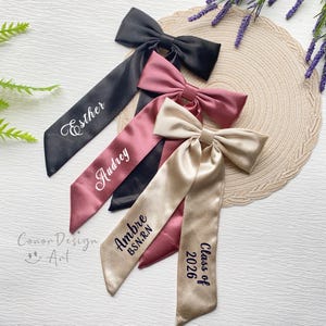 May include: Four satin graduation sashes in black, mauve, and champagne colors. Each sash features a bow and personalized text in white or navy. One sash reads "Esther", another "Audrey", a third "Ambre B.S.N.R.N", and the last "Class of 2026".