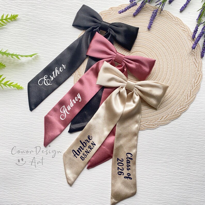 May include: Four satin graduation sashes in black, mauve, and champagne colors. Each sash features a bow and personalized text in white or navy. One sash reads "Esther", another "Audrey", a third "Ambre B.S.N.R.N", and the last "Class of 2026".