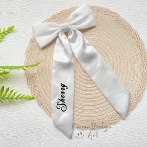 May include: A white satin bow with long streamers. The word "Sherry" is printed in black script on one streamer. The bow is on a beige woven placemat. The words "Conor Design Art" and a smiley face are at the bottom.