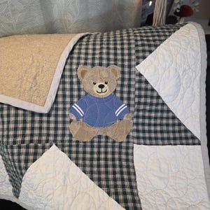 May include: A handmade quilt featuring a teddy bear applique. The quilt has a green and black plaid pattern with white quilted sections. The teddy bear is wearing a blue sweater. The quilt has a beige border.