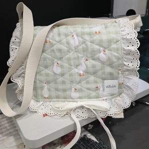 May include: A quilted crossbody bag in light green and white gingham fabric featuring a repeating white goose pattern. The bag is trimmed with white eyelet lace ruffles, a white ribbon bow at the bottom centre, and a woven strap. A small label reads "tiny threads".