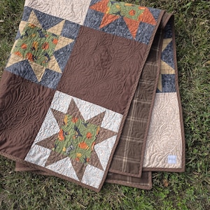 May include: A patchwork quilt featuring star-shaped blocks with green fabric printed with pumpkins and cats, alternating with solid brown blocks quilted with a feather pattern. The quilt includes tan and brown plaid sections and a small white label.