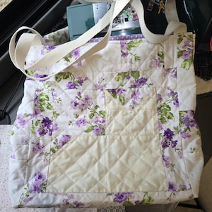 Könnte beinhalten: Eine gesteppte Tragetasche mit weißem Stoffgrund und einem violett-grünen Blumenmuster. Die Tasche verfügt über eine cremefarbene, gesteppte Applikation in Katzenform auf der Vorderseite sowie zwei cremeweiße Stoffträger an der Oberseite.