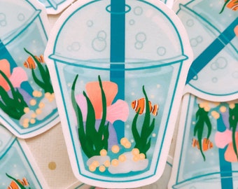 May include: A sticker featuring a cartoon illustration of a bubble tea cup with an underwater scene. The cup is filled with water, coral, plants, and orange and white clownfish. The cup has a teal straw and a clear lid.