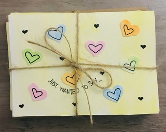 Op de afbeelding: Een stapel gele kaartjes met een strik van bruin touw. De kaartjes zijn versierd met gekleurde papieren hartjes en de tekst "Just wanted to say..." in zwarte stift.