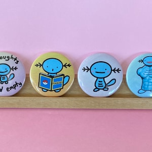 May include: Four round buttons with different designs featuring a blue axolotl character. The first button has a light purple background with the text "no thoughts head empty". The second button has a yellow background with the axolotl holding a book that says "Pokemon". The third button has a light blue background with the axolotl looking straight ahead. The fourth button has a light blue background with the axolotl flexing its muscles.