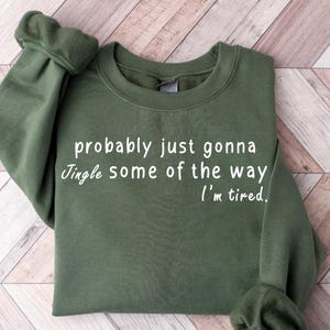 May include: An olive green sweatshirt with the text "probably just gonna jingle some of the way, I'm tired" in white script. The sweatshirt has a crew neck and long sleeves. The garment is made of a soft material.