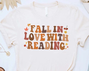 Pode incluir: Uma camiseta branca com o texto "FALL IN LOVE WITH READING" em letras marrons, laranja e vermelhas. O texto é decorado com pequenas imagens de folhas e cerejas.