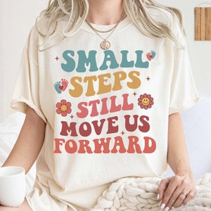 May include: A white t-shirt with the text "SMALL STEPS STILL MOVE US FORWARD" in colourful retro font. The text is decorated with small graphics of footprints and flowers.
