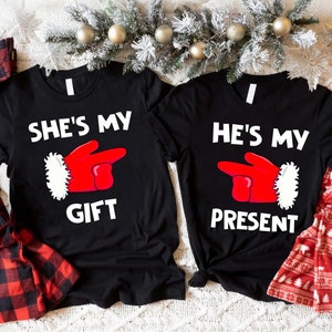 May include: Two black t-shirts with white text and a red and white Santa glove graphic. The left shirt says "She's My Gift" and the right shirt says "He's My Present".