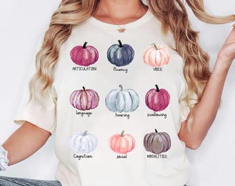 May include: Cream-colored t-shirt featuring nine watercolor pumpkins in various colors. Each pumpkin has a different word written below it, including "Articulation," "Fluency," and "Voice." The shirt is a casual, everyday wear item.