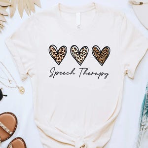 May include: A cream-colored t-shirt with three leopard print hearts and the text "Speech Therapy" in black.