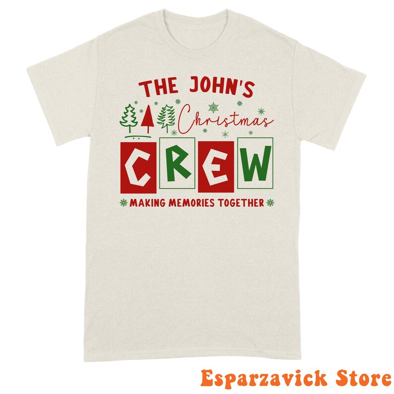 May include: A light beige short-sleeve t-shirt featuring red and green text that reads THE JOHN'S Christmas CREW MAKING MEMORIES TOGETHER. The design includes small green and red pine tree icons and snowflake graphics above the word CREW.