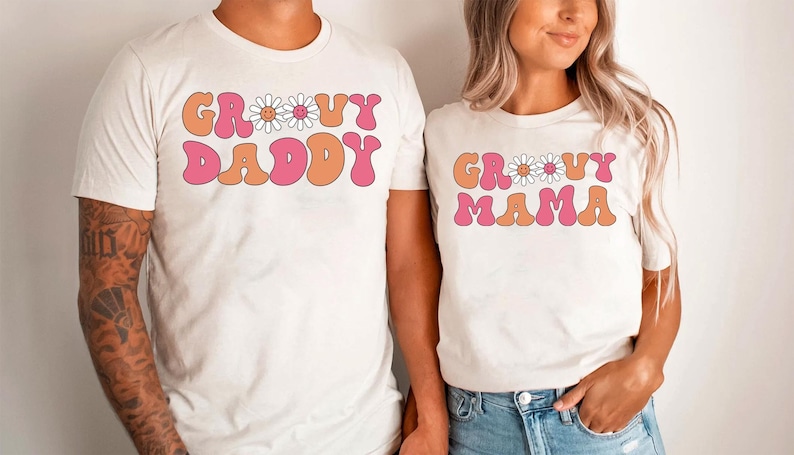 Op de afbeelding: Twee witte T-shirts met een retro lettertype ontwerp. De tekst op de shirts luidt "Groovy Daddy" en "Groovy Mama" met een madeliefje graphic tussen de woorden.