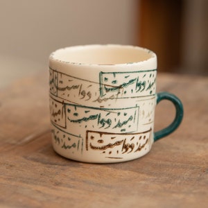 May include: Cream-colored ceramic mug with teal and brown calligraphy. The mug has a teal handle and is sitting on a wooden surface. The calligraphy is in a rectangular frame.