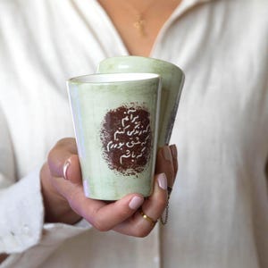 Puede incluir: Un par de tazas cilíndricas de cerámica con un acabado moteado en verde pálido. Cada taza presenta un parche circular de color marrón oscuro en el centro con caligrafía persa blanca impresa en el frente. Superficie exterior lisa.