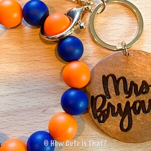 Peut inclure: Un porte-cl&eacute;s en bois avec le texte "Mrs. Bryant" grav&eacute; dessus. Le porte-cl&eacute;s est attach&eacute; &agrave; une boucle perl&eacute;e avec des perles orange et bleues altern&eacute;es.