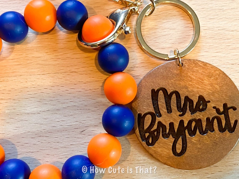 Peut inclure: Un porte-cl&eacute;s en bois avec le texte "Mrs. Bryant" grav&eacute; dessus. Le porte-cl&eacute;s est attach&eacute; &agrave; une boucle perl&eacute;e avec des perles orange et bleues altern&eacute;es.