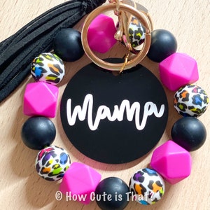 May include: A black and pink beaded keychain with a black circle charm that says "Mama". The keychain has a gold key ring and a black tassel.