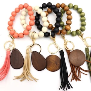 May include: Four wooden beaded bracelets with keychains and tassels. The bracelets are in coral, white, black, and green. The keychains have wooden charms in the shape of a teardrop and a circle. The tassels are in coral, beige, black, and brown.