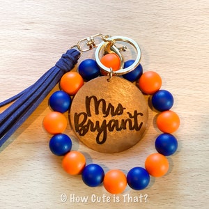 Peut inclure: Un porte-cl&eacute;s en perles bleues et orange avec un charme en bois qui dit "Mme Bryant". Le porte-cl&eacute;s a un anneau de cl&eacute; en or et un pompon bleu.