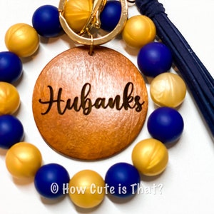 Peut inclure: Un porte-cl&eacute;s en perles dor&eacute;es et bleues avec un charme en bois grav&eacute; du nom "Hubanks".