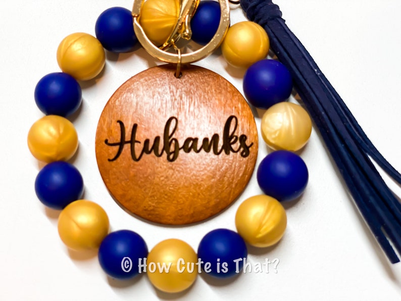 Peut inclure: Un porte-cl&eacute;s en perles dor&eacute;es et bleues avec un charme en bois grav&eacute; du nom "Hubanks".